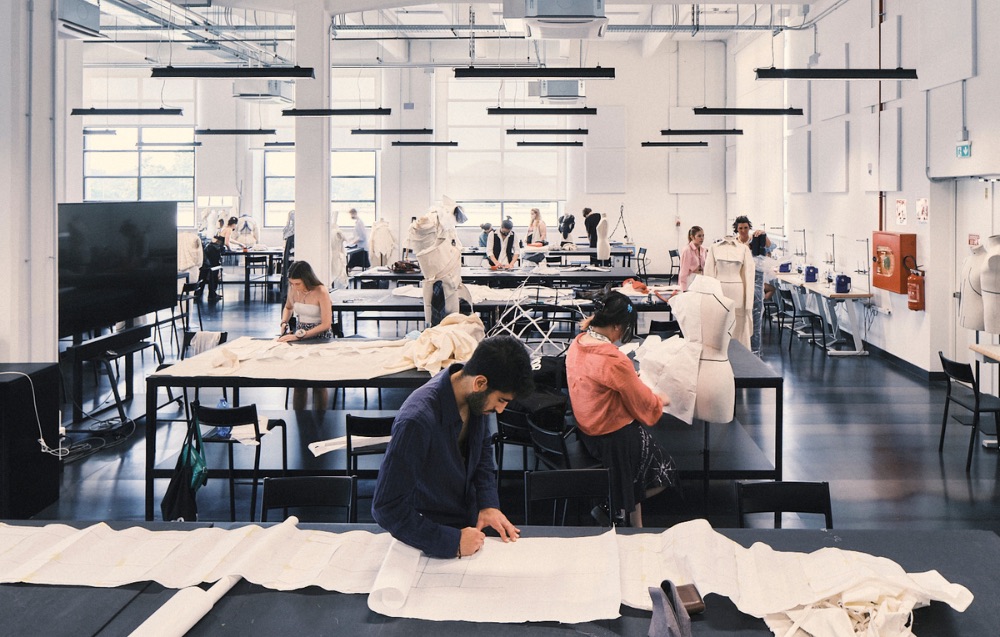 Students at work in a university fashion studio, cutting and draping fabric across long tables beside dress forms.